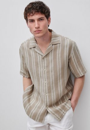 Młody mężczyzna z krótkimi brązowymi włosami, ubrany w beżową i białą koszulę w pionowe paski z krótkim rękawem zapinaną na guziki oraz białe spodnie, ręce w kieszeniach.