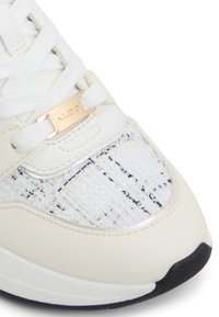 Weiße Sneaker mit strukturiertem Stoff und schwarzen Flecken, glatten Lederakzenten und einer goldfarbigen Logo-Plakette nahe der Schnürsenkel.