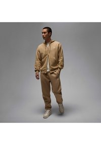 Beige tracksuit set with a zip-up hoodie and tapered pants, featuring a small logo on the chest and thigh. Worn with beige sneakers.