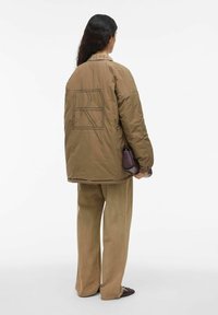 Brun vadderad jacka med stor broderad logga på ryggen, i kombination med beige vida byxor och en liten mörk väska.