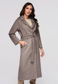 Bruine knie-lange jas gemaakt van een gestructureerde stof. Voorzien van een sjaalkraag, ceintuur in de taille en twee zijzakken. Simpel, strak ontwerp.