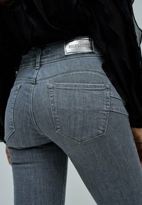 Zbliżenie osoby noszącej dopasowane szare dżinsy z tylnymi kieszeniami i metalową metką "SALSA JEANS" na pasku, w połączeniu z czarną bluzką.