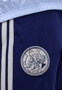 Σκούρο μπλε αθλητικό παντελόνι με δύο ανοιχτές κάθετες ρίγες και κυκλικό σήμα Ajax που απεικονίζει το προφίλ ενός πολεμιστή.