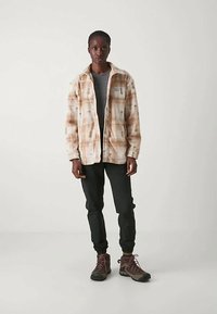 Άτομο που στέκεται φορώντας μια μπεζ και λευκή καρό ζακέτα, σκούρο γκρι πουκάμισο, μαύρα jogger παντελόνια και καφέ μποτάκια πεζοπορίας σε απλό φόντο.