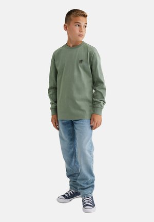 T-shirt verde a maniche lunghe in cotone con un piccolo logo, abbinato a jeans in denim azzurro chiaro e sneakers navy con suole bianche.