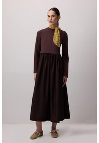 Brown long-sleeve dress with a gathered waist and solid color. A patterned yellow scarf is tied at the neck. Soft fabric and relaxed fit.