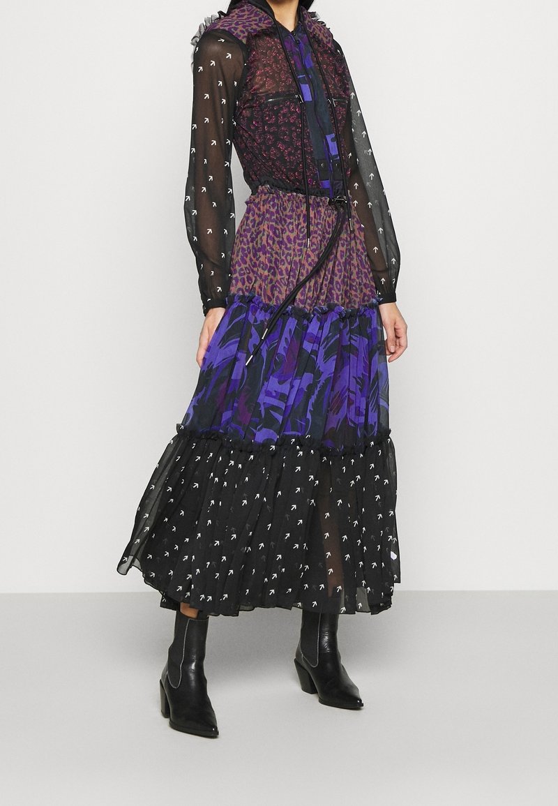 Femme portant une robe longue à volants avec des manches en voile noir, motifs léopard et floraux en violet et noir, associée à des bottes noires à talons.