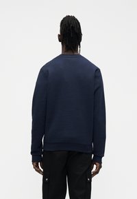 Μπλε σκούρο φούτερ με λαιμόκοψη crew, υφή στο ύφασμα, ριμπ μανσέτες και τελείωμα. Φορεμένο με μαύρο παντελόνι cargo· απλό φόντο.