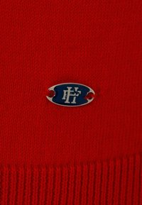Tejido de punto rojo con un logo ovalado en plata y azul, con pequeños agujeros para sujeción, que muestra un patrón texturizado.