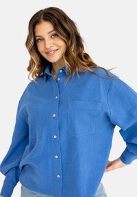 Niebieska koszula z guzikami, wykonana z teksturowanej tkaniny, z dwiema kieszeniami na piersi, kołnierzykiem oraz lekko oversize'owymi rękawami.
