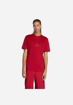 Junger Mann mit geflochtenem Haar, der ein rotes Adidas-T-Shirt und passende rote Shorts trägt, steht vor einem schlichten hellen Hintergrund.