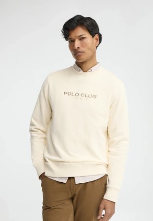Uomo che indossa una felpa color crema con la scritta "POLO CLUB", camicia beige sotto e pantaloni marroni, in piedi davanti a uno sfondo semplice.