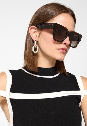Donna con capelli castani lisci indossa grandi occhiali da sole tartarugati, orecchini a catena d'argento e un top nero senza maniche con bordi bianchi.