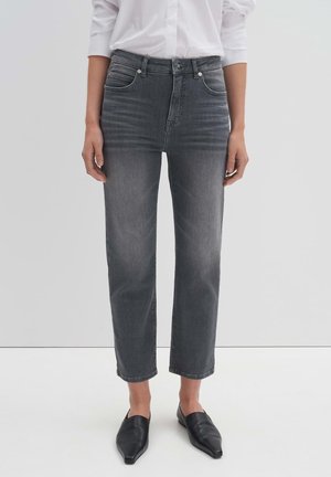 Person iført grå jeans med høj talje og lige ben, hvid skjorte stoppet ind og sorte flade sko med spids snude, stående på hvidt gulv.