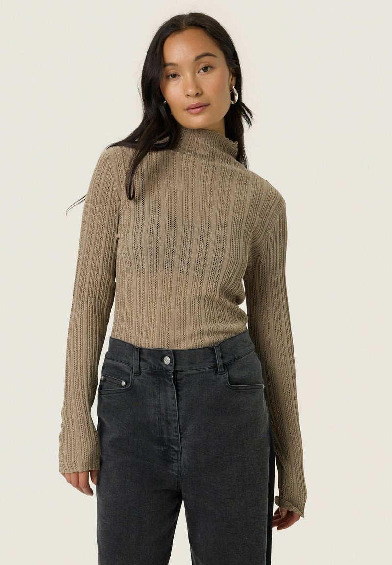 Ung kvinde med langt, mørkt hår iført en ribbet, beige turtleneck sweater og højtholdsende sorte jeans, stående foran en ensfarvet lys baggrund.