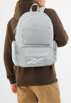 Person trägt einen hellgrauen Reebok-Rucksack mit mehreren Reißverschlussfächern über einem braunen Hoodie und hellbraunen Hosen.
