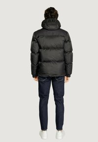 Giacca trapuntata nera con colletto alto e logo in rilievo, polsini a costine, abbinata a jeans in denim scuro e sneaker bianche.