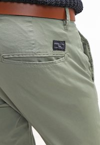 Ljusgröna byxor med en mjuk yta, en bakficka med en svart etikett, och ett brunt bälte som är bärt i midjan.