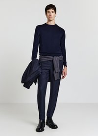 Marineblauer Pullover, graue Hose mit strukturiertem Muster und schwarze Chelsea-Stiefel. Eine passende graue Jacke ist lässig um die Taille gelegt.