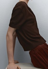 Brun viltt kortärmad cardigan med rund hals och knappar, tillsammans med lösa röda byxor, placerad mot en beige yta.
