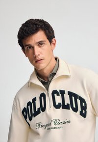 Pullover in pile color crema con colletto a mezza zip, caratterizzato da ricami verdi con la scritta "POLO CLUB" e "Beyond Classics". Texture morbida.