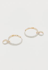 Χρυσά σκουλαρίκια κρίκοι με μικρούς και μεγάλους κρίκους διακοσμημένους με διάφανα στρας σε λευκό φόντο.