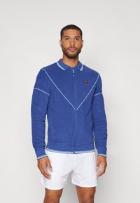 Blå zip-up hettegenser med krage, hvite pipingdetaljer, ribbede mansjetter og en liten logo på venstre bryst. Stoff av bomullsblanding.
