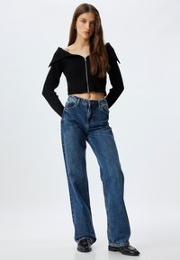 Czarny prążkowany crop top z zamkiem z przodu i szerokim kołnierzem, zestawiony z niebieskimi dżinsami o wysokim stanie. Modelka nosi czarne buty.