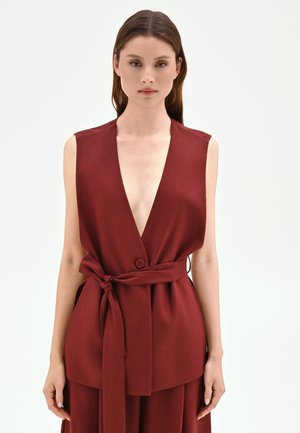 Femme portant un gilet bordeaux sans manches à décolleté en V profond avec une ceinture en tissu nouée à la taille, debout devant un fond blanc uni.
