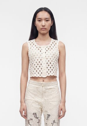 Femme aux longs cheveux noirs portant un crop top blanc en crochet sans manches et un pantalon en dentelle blanche taille haute, debout devant un fond uni.