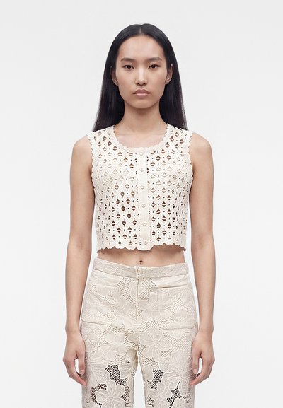 Femme aux longs cheveux noirs portant un crop top blanc en crochet sans manches et un pantalon en dentelle blanche taille haute, debout devant un fond uni.