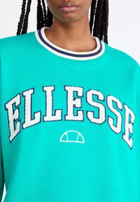 Persona con sudadera verde azulado con cuello a rayas blancas y azul marino y grandes letras blancas "ELLESSE" bordadas en el frente.