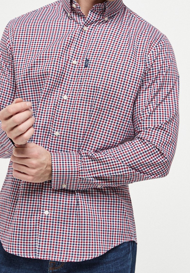 Homme ajustant le poignet d'une chemise à petits carreaux rouges, blancs et bleus boutonnée, portant un jean bleu, sur fond blanc.