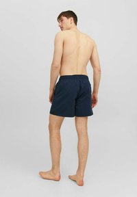 Costume da bagno blu navy con vita elasticizzata e tasche laterali, caratterizzato da una texture liscia e un design semplice. Indossato da una persona vista da dietro.