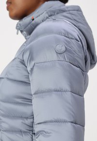 Giacca a vento leggera di colore blu chiaro con cappuccio, con sezioni trapuntate, texture lucida e un logo circolare sulla spalla sinistra.