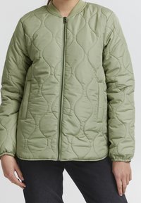  Pikowana kurtka bomberka w jasnym zielonym kolorze, z przednim zamkiem błyskawicznym, żebrowanym kołnierzem oraz bocznymi kieszeniami i elastycznymi mankietami dla lepszego dopasowania.