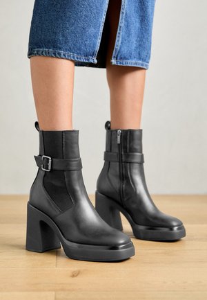 Bottines noires en cuir avec un talon bloc haut, des panneaux latéraux élastiques et une boucle décorative. Texture lisse avec un design élégant.