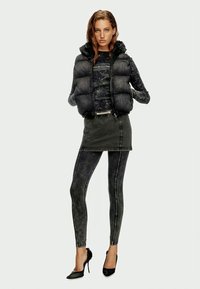 Schwarze Steppweste über einem schwarzen gemusterten Oberteil, kombiniert mit einem kurzen schwarzen Jeansrock und figurbetonten schwarzen Leggings, akzentuiert mit hohen Absätzen.