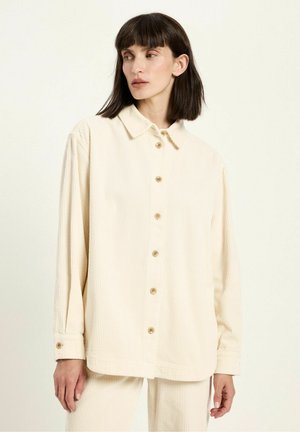 Woman with dark hair wearing an oversized cream corduroy button-up shirt and matching pants against a plain light background.