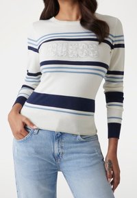 Suéter color crema claro con rayas horizontales en azul marino y azul, con un logo "GUESS" en detalle texturizado en el frente. Diseño de ajuste slim.