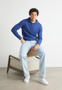 Felpa con cappuccio blu e un piccolo logo, indossata con jeans azzurri e sneakers bianche, seduto su una sedia di legno con un sedile imbottito.