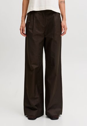 Personne portant un pantalon large marron foncé et un haut blanc, debout devant un fond uni gris clair.