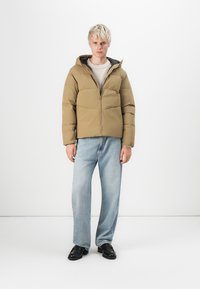 Brown puffer jacket with a front zipper and hood, featuring a rectangular quilted design. Worn with light blue jeans and black shoes.