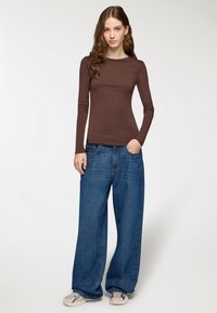 Giovane donna in piedi con una mano in tasca, indossando un maglione marrone a maniche lunghe, jeans larghi blu e sneaker beige su sfondo bianco.