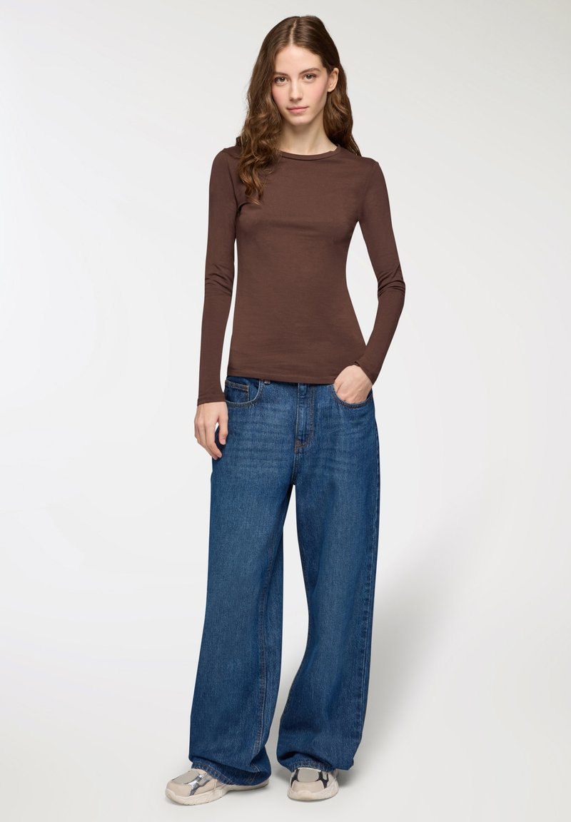 Giovane donna in piedi con una mano in tasca, indossando un maglione marrone a maniche lunghe, jeans larghi blu e sneaker beige su sfondo bianco.