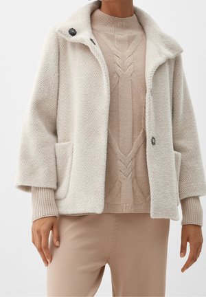 Nainen pukeutunut kermanväriseen kalakuvioiseen villatakkiin, jonka alla on beige palmikkoneule ja siihen sopivat beiget housut, seisoo valkoista taustaa vasten.