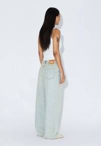 Jean bleu clair à jambes larges en denim, avec un design simple et des coutures discrètes. L'étiquette arrière est orange avec le nom de la marque.