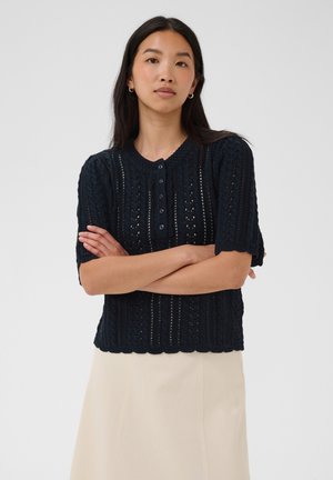Frau mit langen dunklen Haaren, die einen kurzärmligen marineblauen Strickpullover und einen hellbeigen Rock trägt, steht mit verschränkten Armen.
