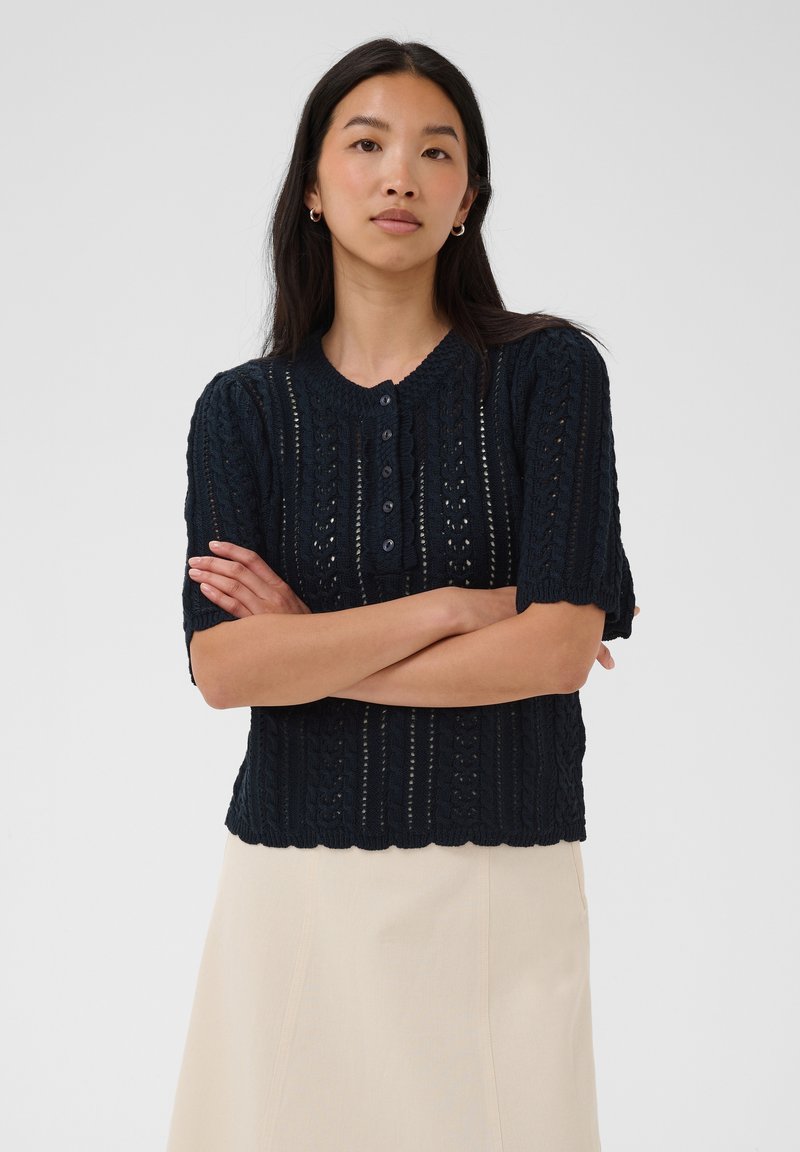 Femme aux longs cheveux foncés portant un pull en maille bleu marine à manches courtes et une jupe beige clair, debout les bras croisés.