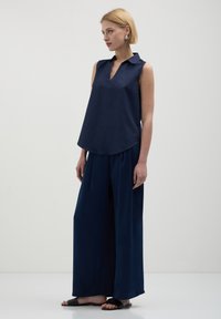 Donna che indossa un top collare senza maniche di colore blu scuro e pantaloni a gamba larga, in piedi a piedi nudi su uno sfondo chiaro e semplice.